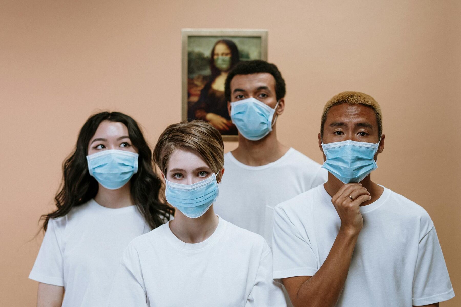 A group of four people wearing white t-shirts and blue face masks, standing in front of a painting