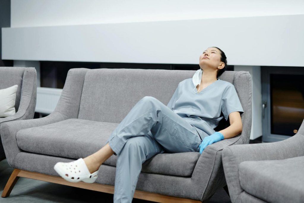 A nurse in scrubs resting on a sofa with her eyes closed, taking a break in a modern healthcare setting