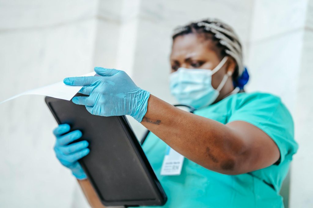 A nurse in scrubs reviews a document while wearing gloves and a mask
