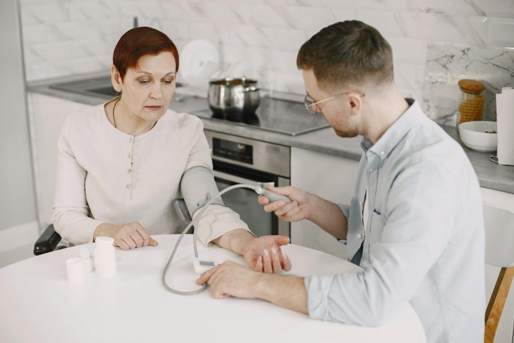 A caregiver checks an elderly woman's blood pressure at a kitchen table