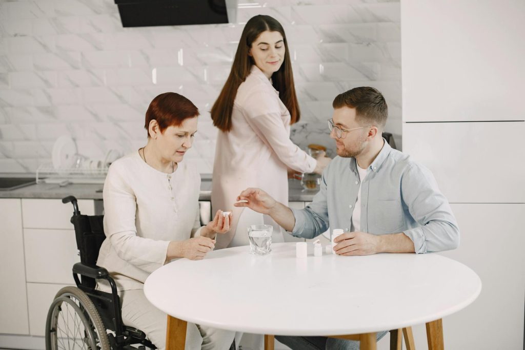 An elder woman in a wheelchair is seated at a table, engaged in conversation with a man and a woman beside her