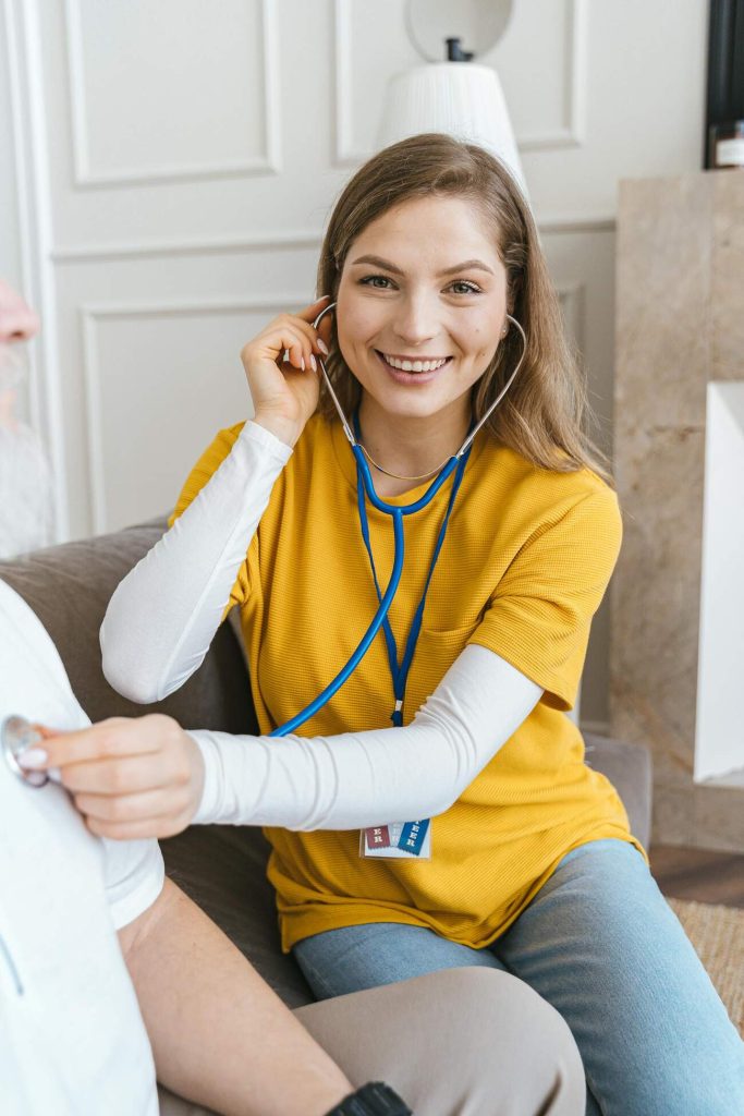 A woman sits on a couch, wearing a stethoscope around her neck, checking a persons heartbeat