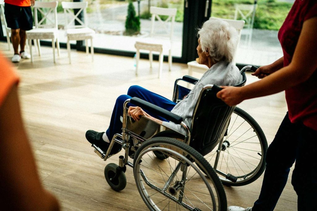 An elderly person being pushed in a wheelchair by a caregiver in a brightly lit room